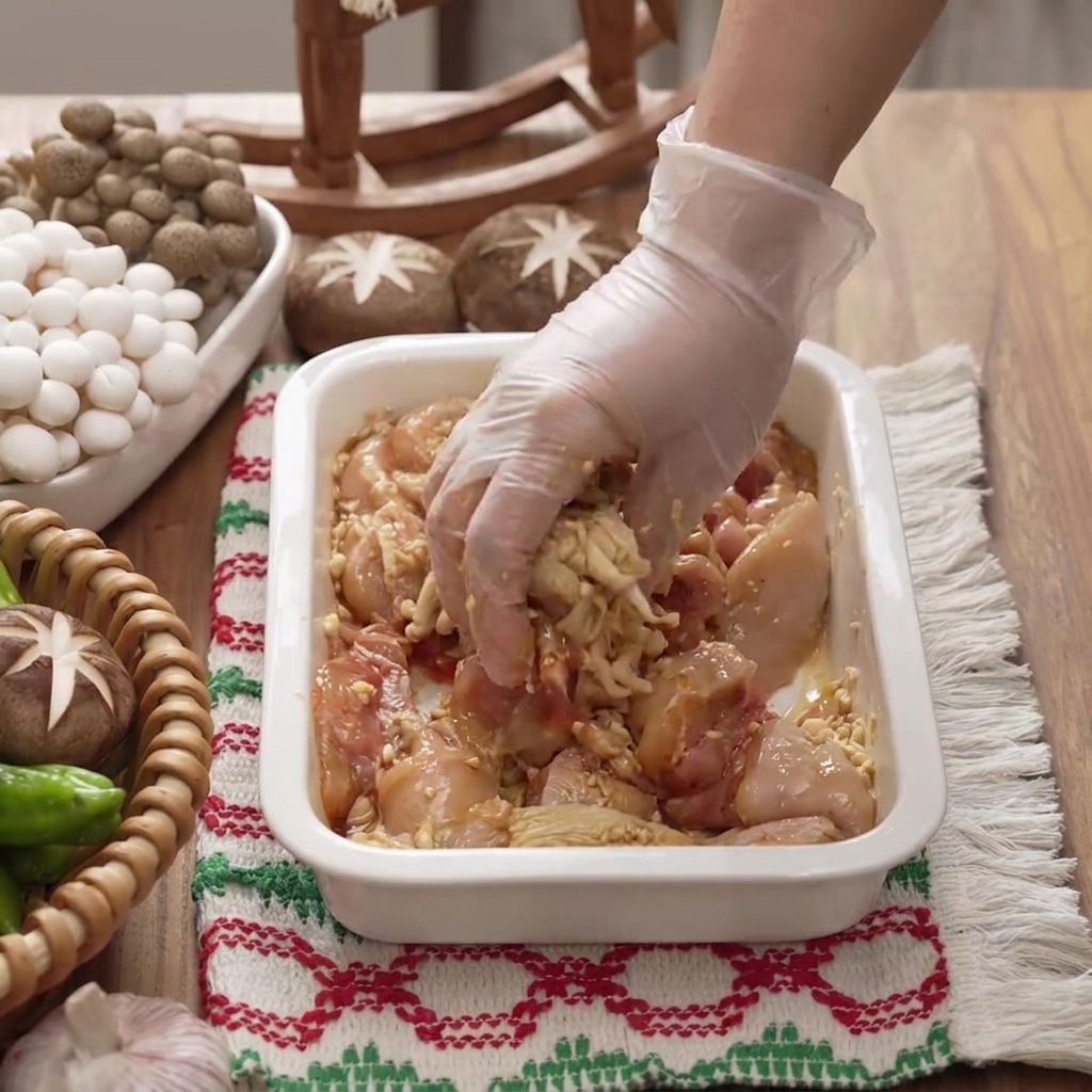 Eine handschuhte Hand mischt und massiert Marinade in Hähnchenstücke in einer weißen Schale.