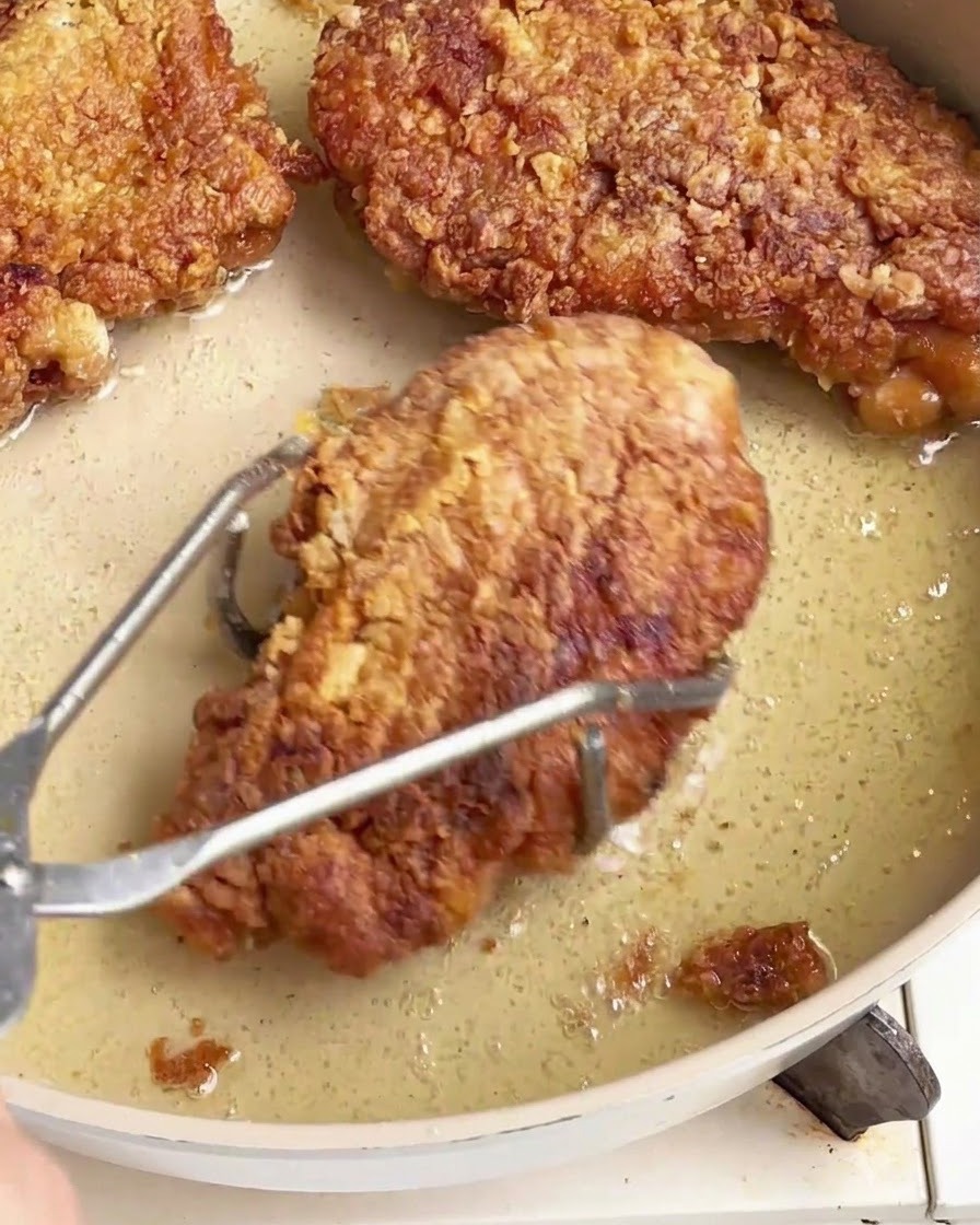 Knusprig frittierte Hähnchenstücke werden mit einer Edelstahlzange aus einer öligen Pfanne gehoben.