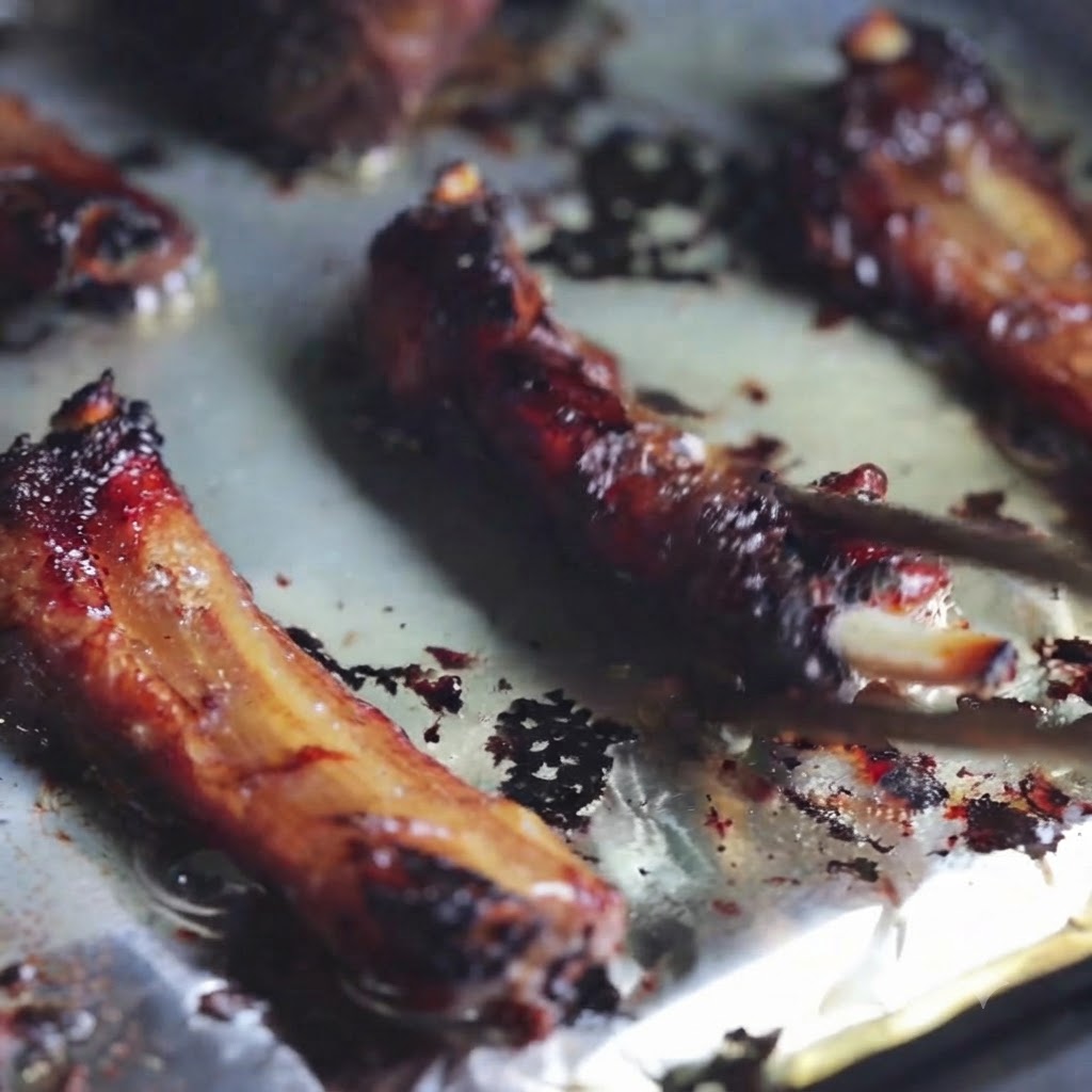 Nahaufnahme von gegrillten Schweinerippchen, die mit Stäbchen auf einem mit Folie ausgelegten Backblech umgedreht werden.