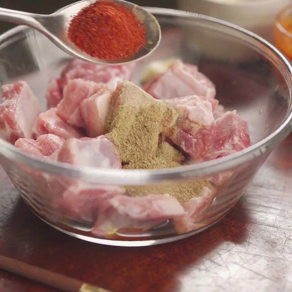 Rotes Chilipulver auf einem Löffel über einer Schüssel mit rohen Schweinerippchen, bedeckt mit Gewürzen und Kräutern.