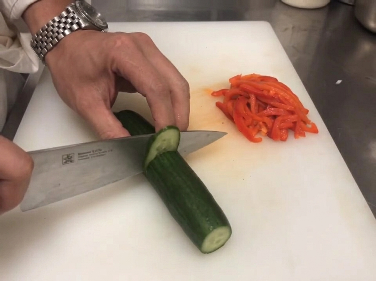 Ein Messer schneidet eine frische grüne Salatgurke auf einem Brett neben roten Paprikastreifen in feine Stifte.