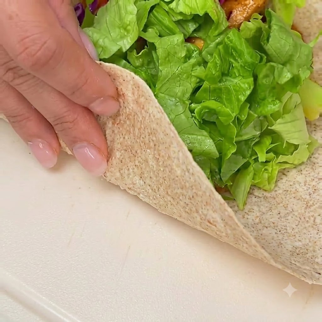 Hände falten und rollen eine gefüllte Vollkuorn-Tortilla auf einer sauberen weißen Oberfläche.