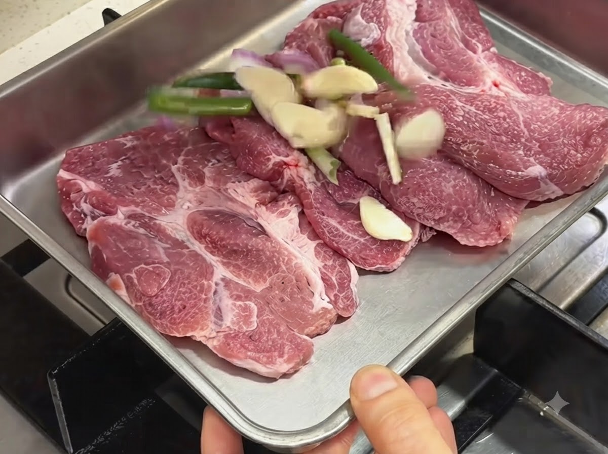 Rohe Schweinekoteletts in einer Metallschale neben geschnittenem Knoblauch, Frühlingszwiebeln und Schalotten.