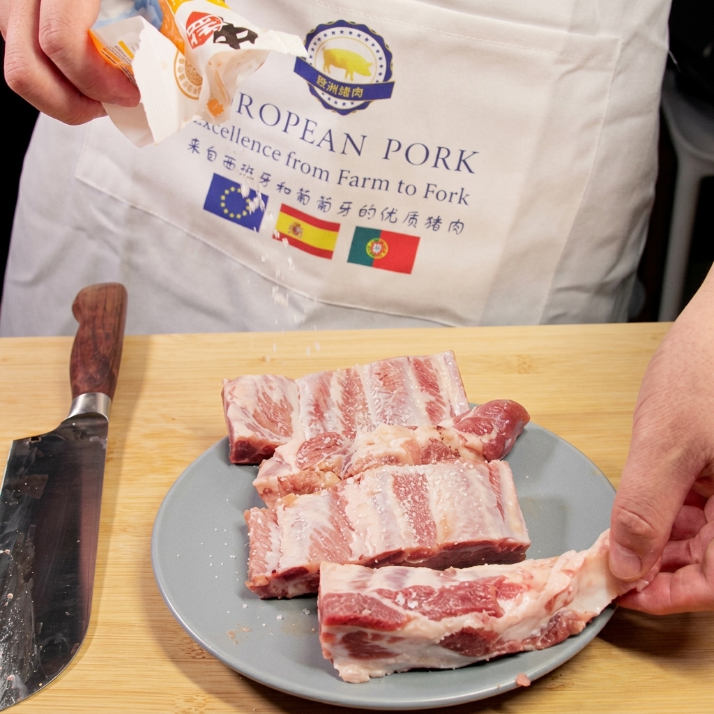 Salz wird aus einer Verpackung auf rohe Schweinerippchen gestreut, die auf einem Teller neben einem Chefmesser liegen.