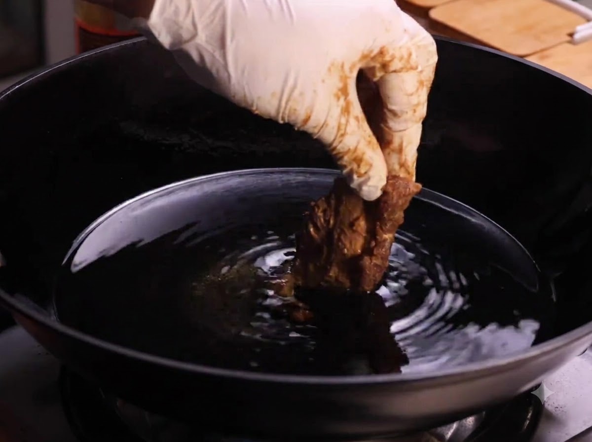 Eine behandschuhte Hand senkt ein Stück paniertes Schweinerippchen in einen mit heißem Speiseöl gefüllten Wok.