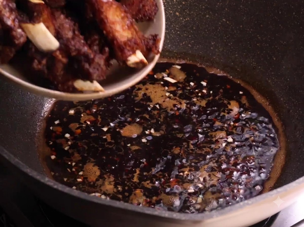 Frittierte Schweinerippchen werden von einem Teller in die köchelnde dunkle Sauce in einem Wok gegeben.