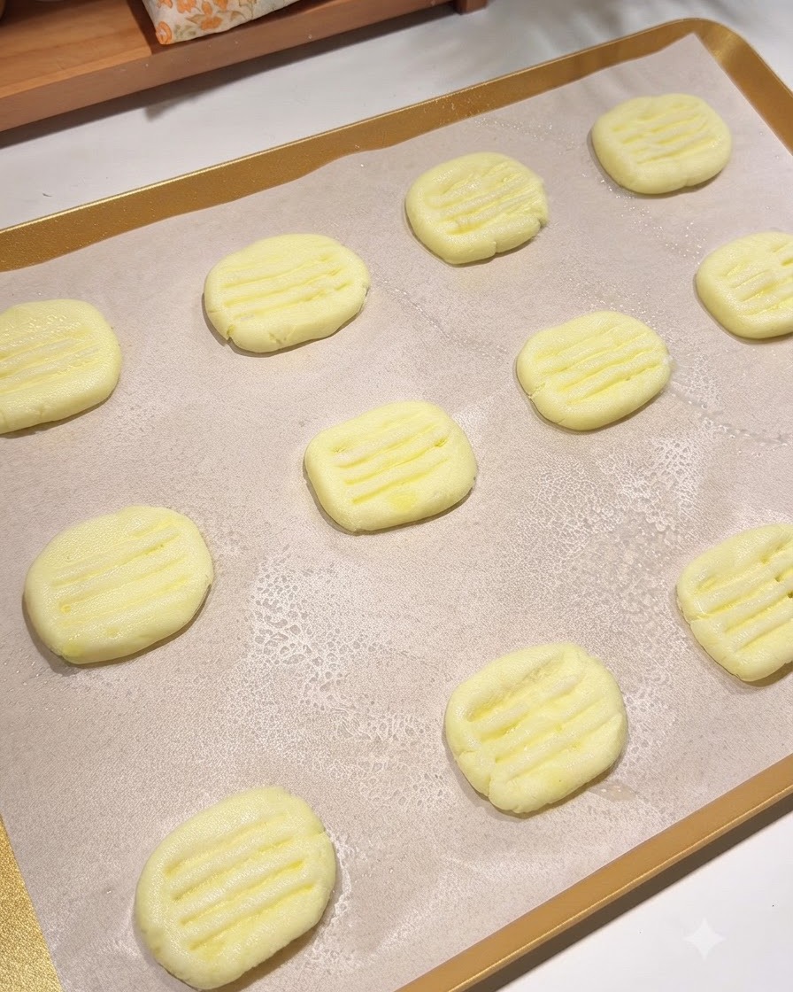 Ein mit Backpapier ausgelegtes Backblech mit mehreren geformten Kartoffelpuffern, auf die feiner Sprühnebel aus Speiseöl gesprüht wird.