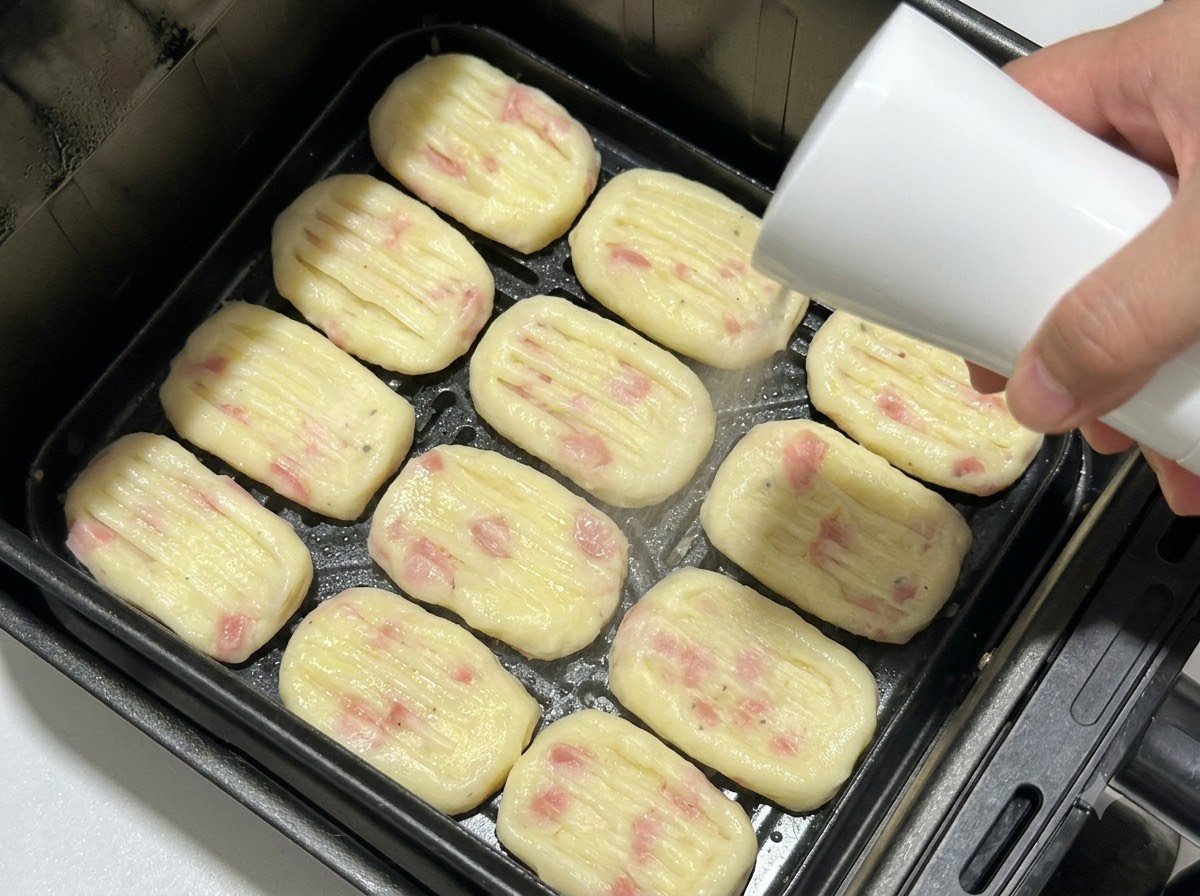 Geformte Kartoffeltaler in einem Heißluftfritteusen-Korb angeordnet, während sie mit einer Sprühflasche mit Öl besprüht werden.