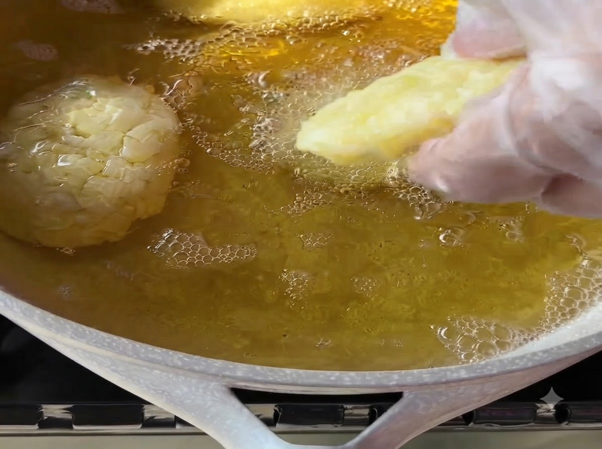 Eine behandschuhte Hand legt vorsichtig einen geformten Kartoffeltaler in eine Pfanne mit heißem, blubberndem Frittieröl.