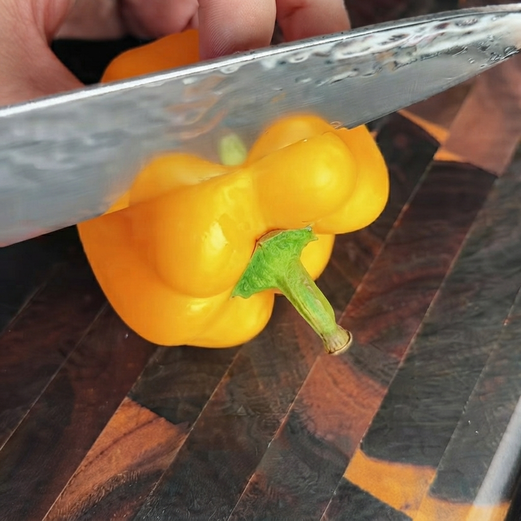 Ein scharfes Messer schneidet den oberen Teil einer hellgelben Paprikaschote auf einem Holzschneidebrett ab.