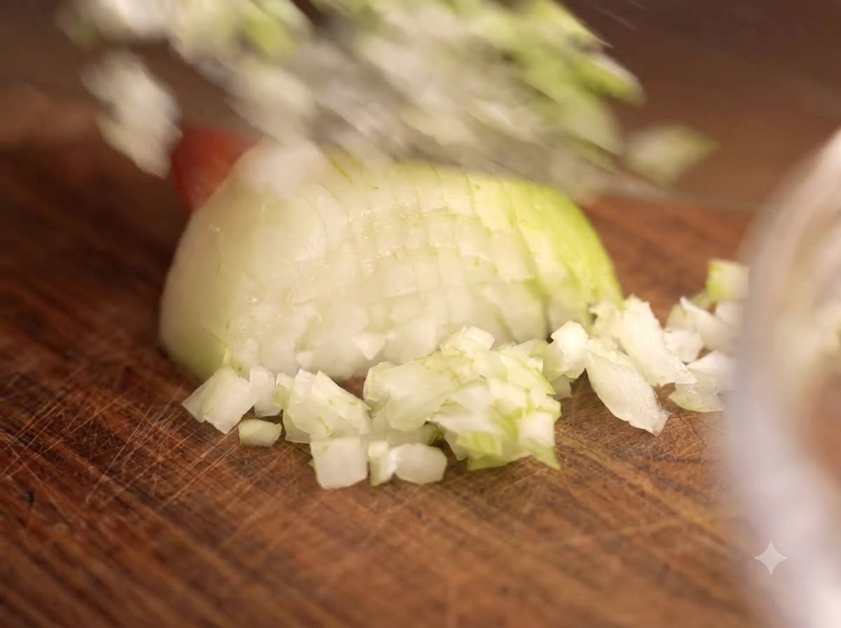 Ein Messer zerkleinert Zwiebeln und Knoblauch auf einem Holzbrett neben einer Glasschüssel mit Fleisch.
