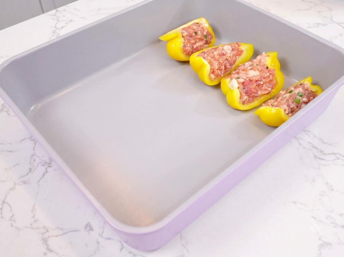Gelbe Paprikahälften mit gewürztem Hackfleisch ordentlich in einer lila Backform angeordnet.