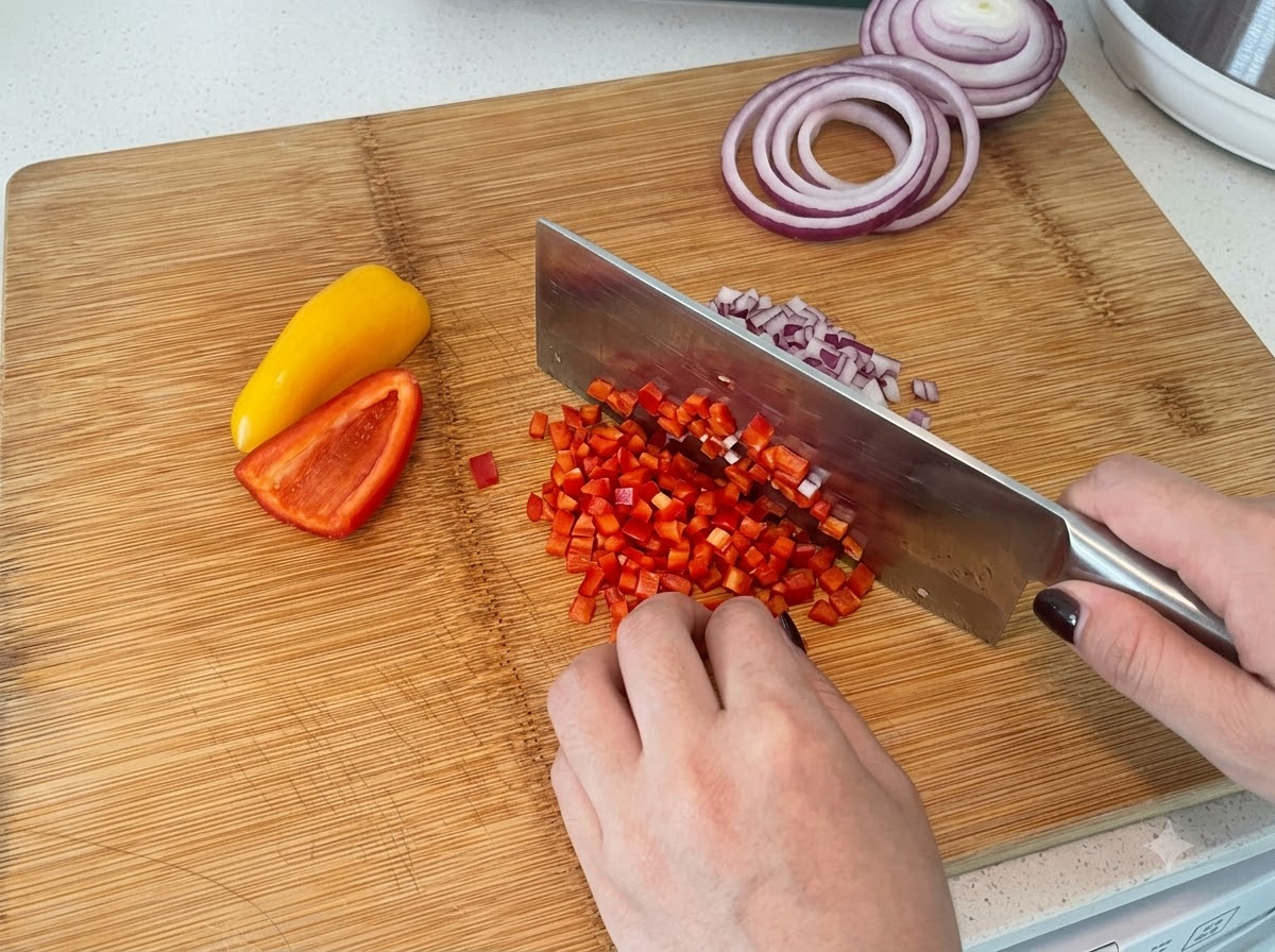 Rote und gelbe Paprika werden auf einem Holzschneidebrett mit einem Küchenbeil in kleine Würfel geschnitten.