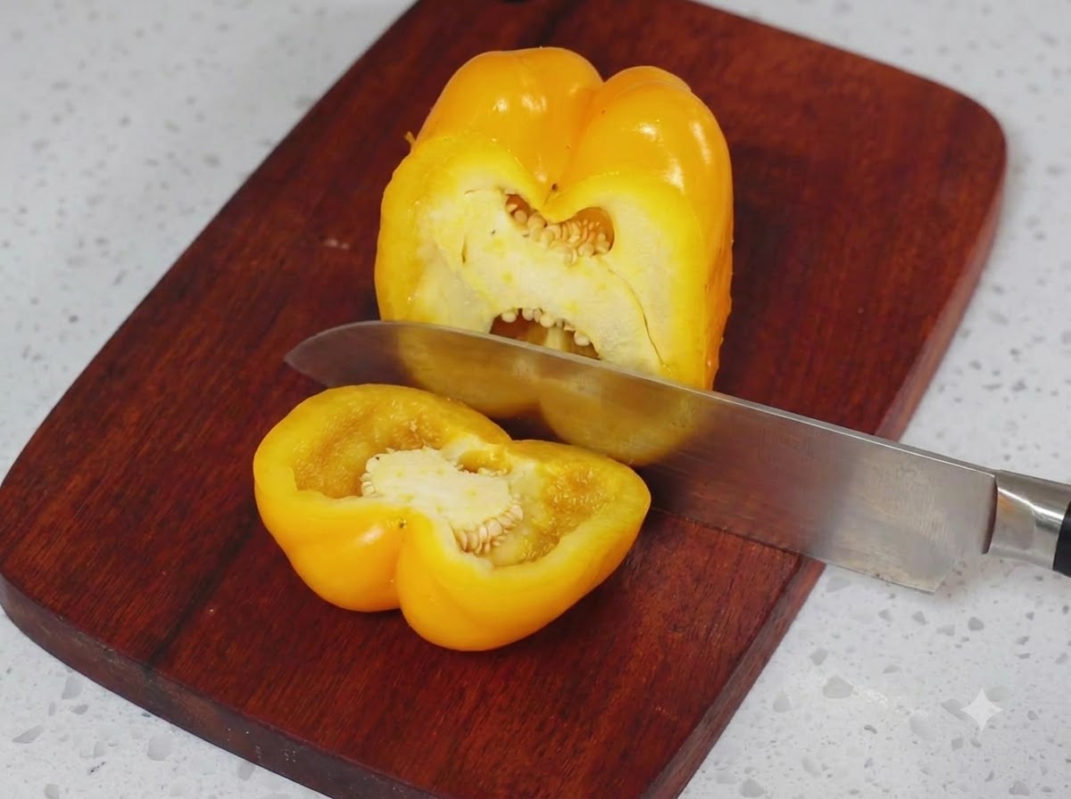 Eine gelbe Paprika wird auf einem Holzschneidebrett mit einem Küchenmesser in der Hälfte geschnitten.