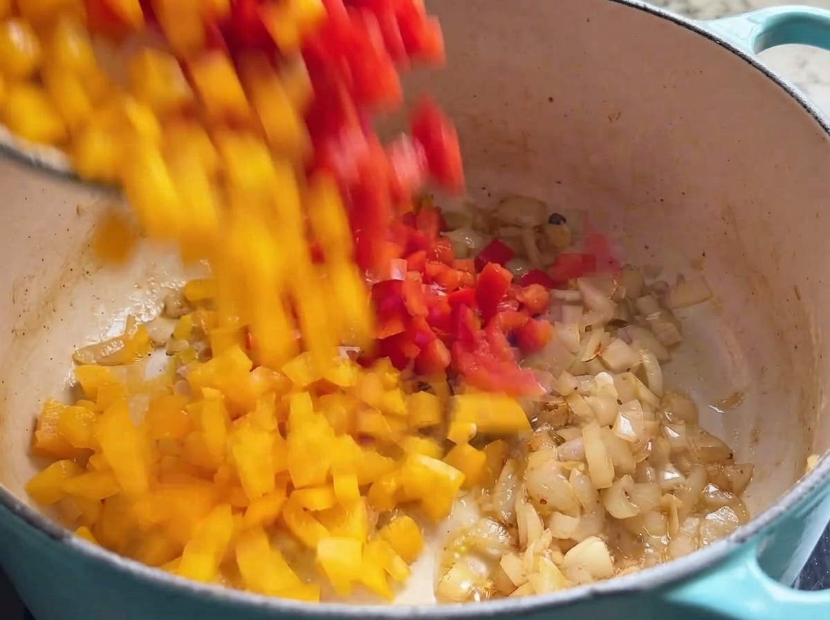Frisch gewürfelte rote und gelbe Paprikastücke fallen in einen Kochtopf gefüllt mit angeschwitzten Zwiebeln.