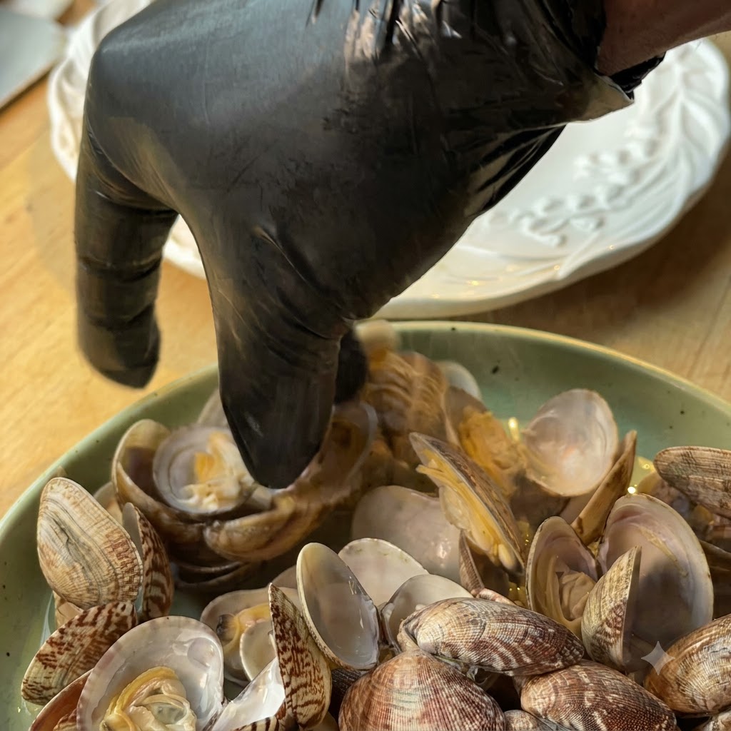 Eine Person mit schwarzen Handschuhen pflückt Muschelfleisch aus den Schalen auf einen Teller.
