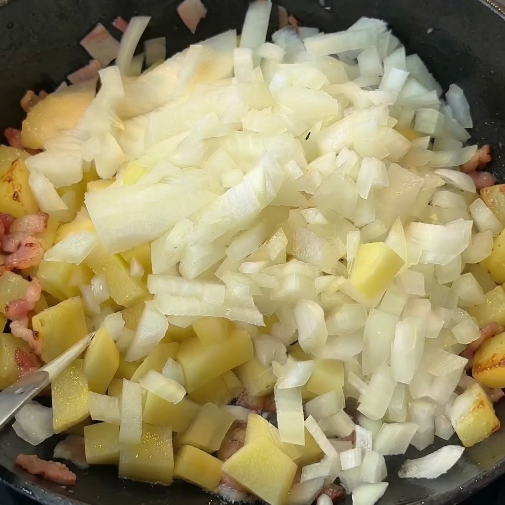 Gewürfelte Zwiebeln werden in einer Pfanne auf gekochte Kartoffeln und Speck gegeben.
