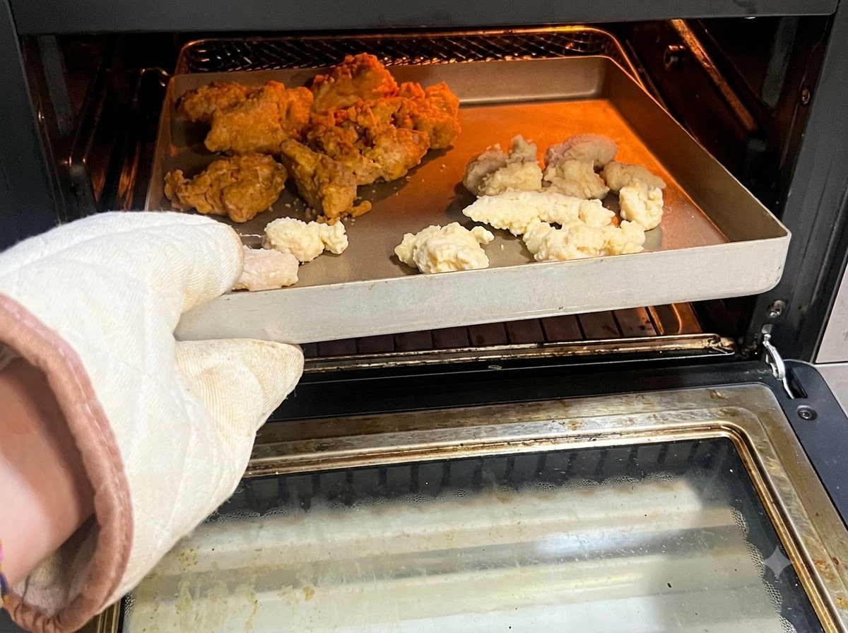 Ein Ofenhandschuh hält einen Korb gefüllt mit rohem oder tiefgekühltem Popcorn-Hähnchen und scharfen Flügeln in einer Heißluftfritteuse.