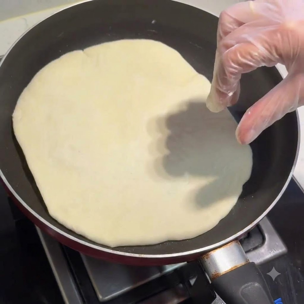 Ein Fladenbrot backt in einer Bratpfanne, während eine behandschuhte Hand es anpasst.