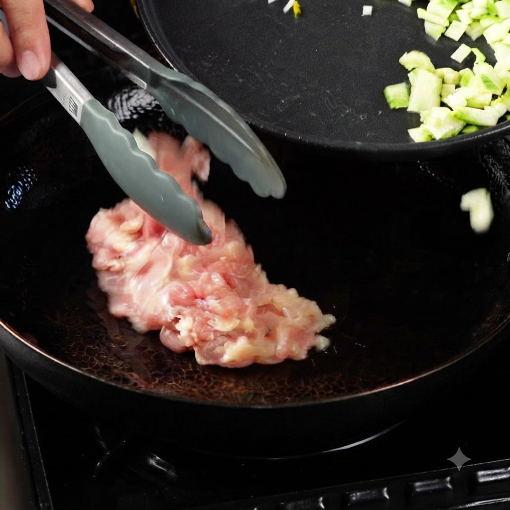 Zange legt rohe Hähnchenstücke in einen erhitzten Wok mit Öl, neben einer Pfanne mit gewürfeltem Gemüse.