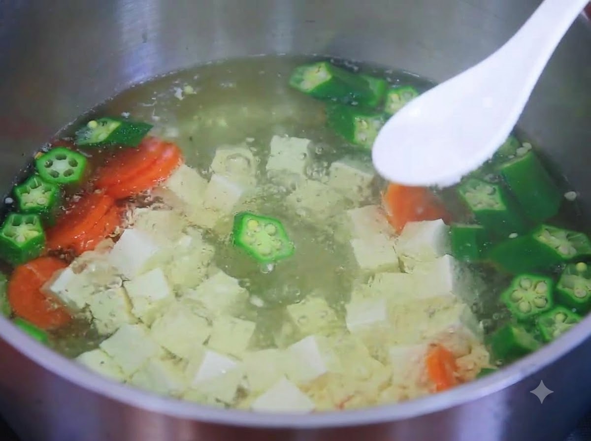 Ein weißer Löffel hält Salz über einen köchelnden Topf mit Tofu-, Okra- und Karottensuppe.
