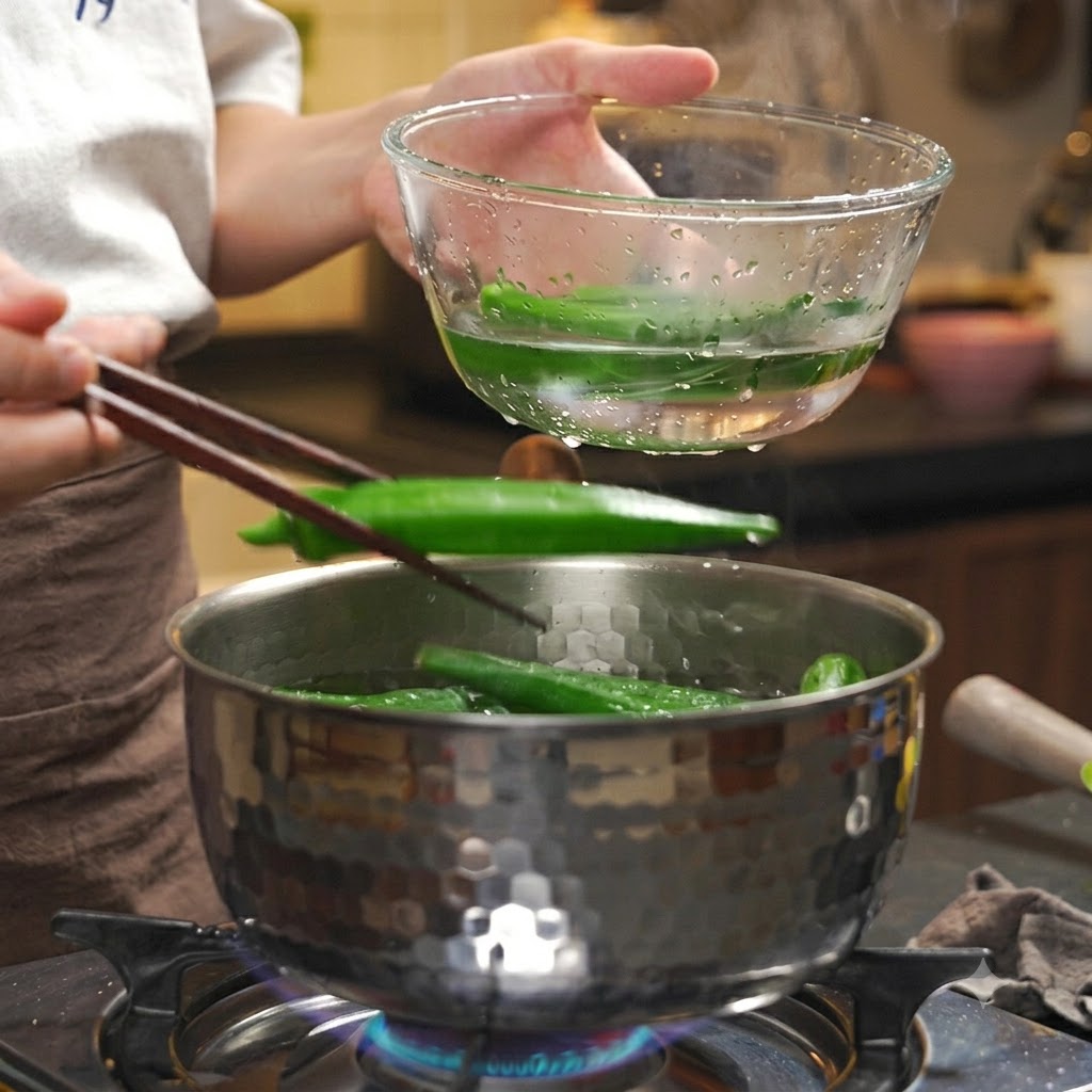 Gekochte Okra wird mit Essstäbchen aus einem heißen Topf in eine klare Schüssel mit Eiswasser gegeben.
