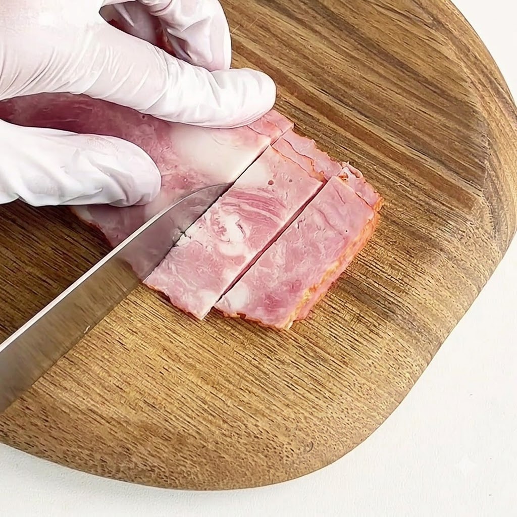 Eine handgeschützte Hand schneidet gepökeltem Speck auf einem runden Holzschneidebrett in kleine Stücke.