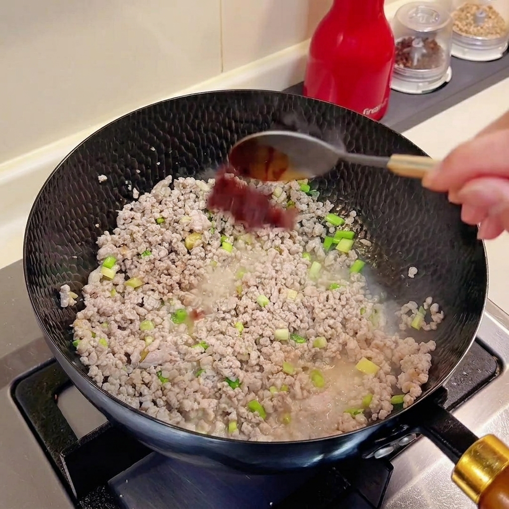 Ein Löffel Doubanjiang wird in einen Wok mit Hackfleisch und Gewürzen gegeben.