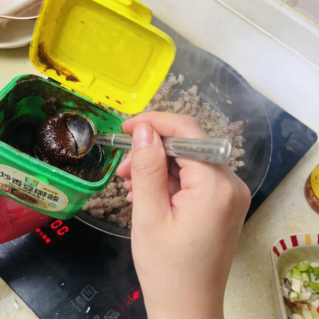 Ein Löffel dunkle Bohnenpaste wird aus einem grünen Behälter in das angebratene Schweinegehacktes geschöpft.
