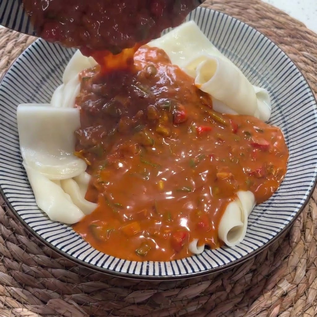 Eine reichhaltige rote Sauce wird aus einer Kelle auf eine Schüssel mit gefalteten breiten Nudeln gegossen.