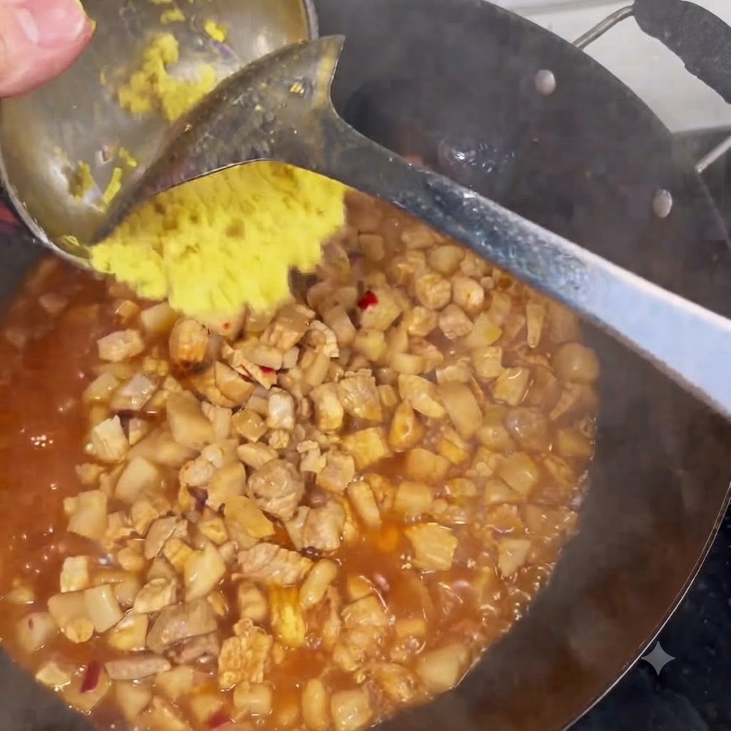 Gehackter Ingwer wird von einer Kelle in einen Wok geschabt, der mit schmorendem Fleisch und Soße gefüllt ist.