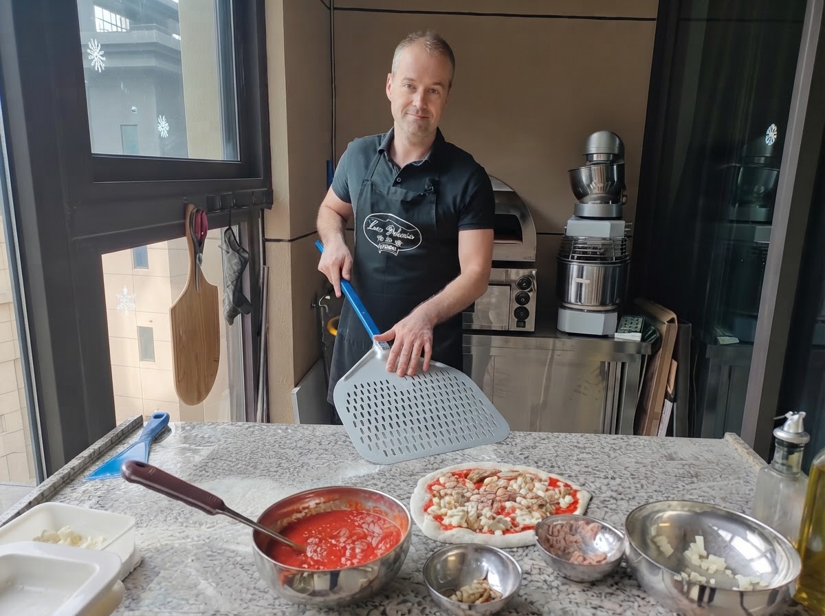 Ein Pizzabäcker hält einen großen gelochten Pizzaschieber aus Metall direkt neben einer fertig belegten rohen Pizza.