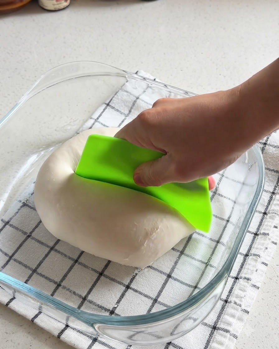 Eine Hand schneidet mit einer hellgrünen Teigkarte eine glatte Pizzateigkugel in einer Glasschüssel in zwei Hälften.