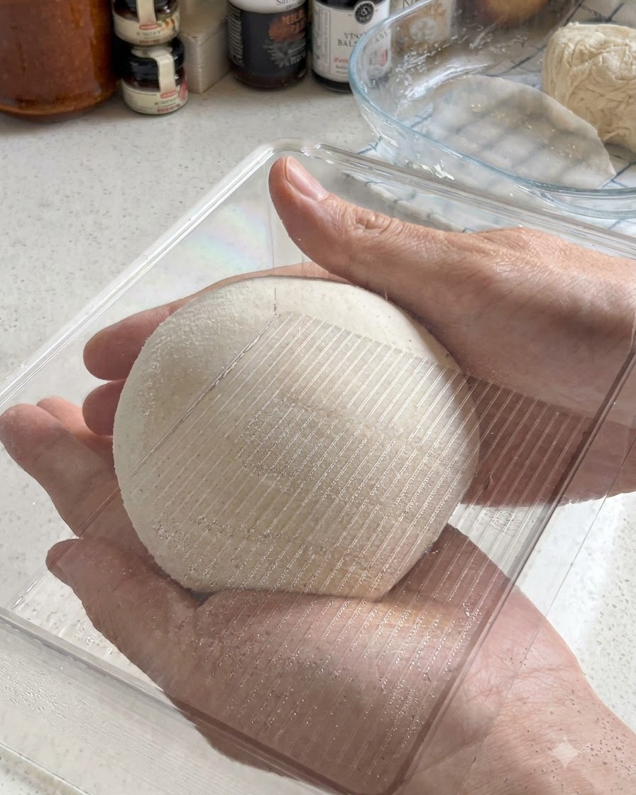 Zwei Hände halten eine perfekt abgerundete Pizzateigkugel, während sie in eine transparente Plastik-Gärbox gelegt wird.