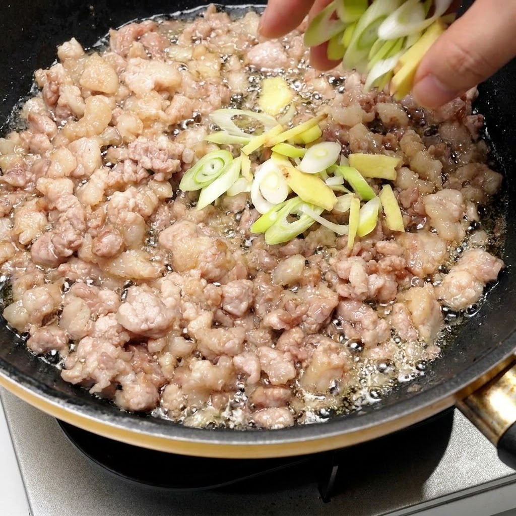 Gehackte Frühlingszwiebeln und Ingwer werden direkt in die Pfanne mit dem gebräunten Schweinefleisch gegeben.