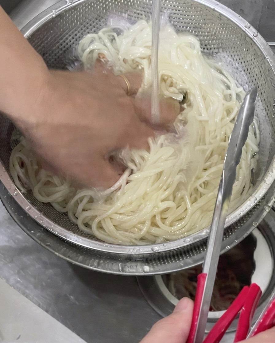 Eine Hand schwenkt gekochte Udon-Nudeln in einem Metallsieb unter kaltem Leitungswasser.