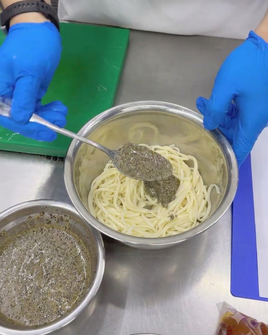 Mit Handschuhen versehene Hände geben mithilfe eines Löffels dicke Trüffelsauce zu kalten Udon-Nudeln in einer Rührschüssel.