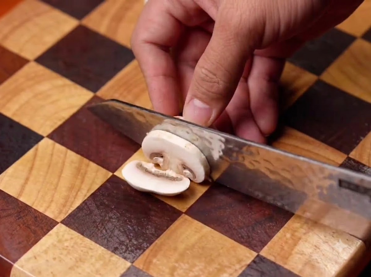 Champignons werden mit einem Messer auf einem karierten Holzschneidebrett geschnitten und anschließend in eine schwarze Pfanne gegeben.