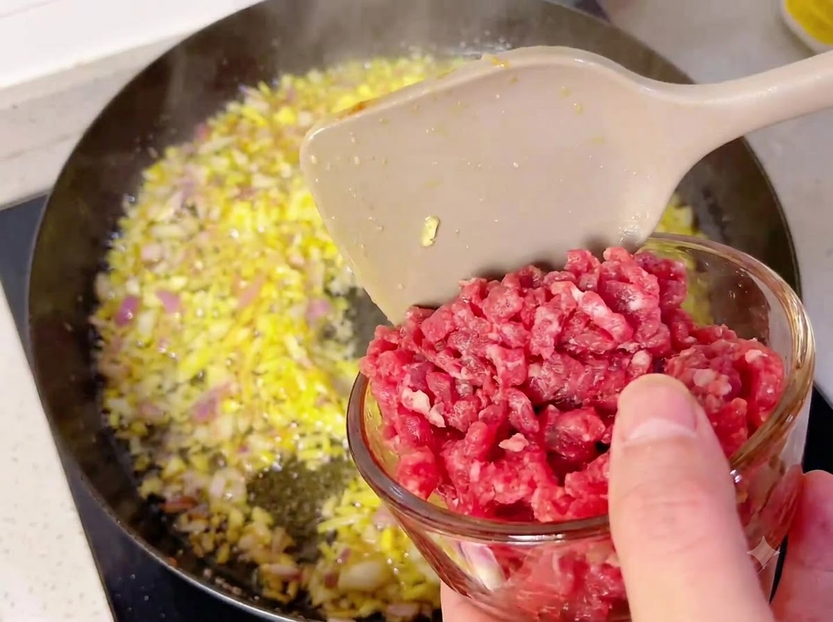 Eine kleine Glasschüssel gibt Rindfleischwürfel in eine heiße Pfanne mit angeschwitzten Zwiebeln und Knoblauch.