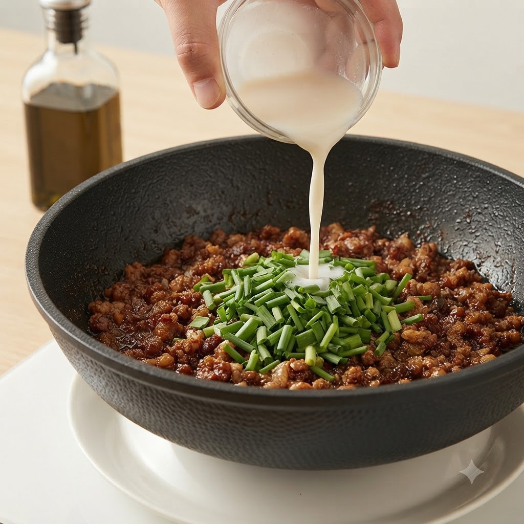 Eine Hand gießt eine weiße Stärkemischung aus einer kleinen Glasschüssel in eine Pfanne mit Hackfleisch und Schnittlauch.
