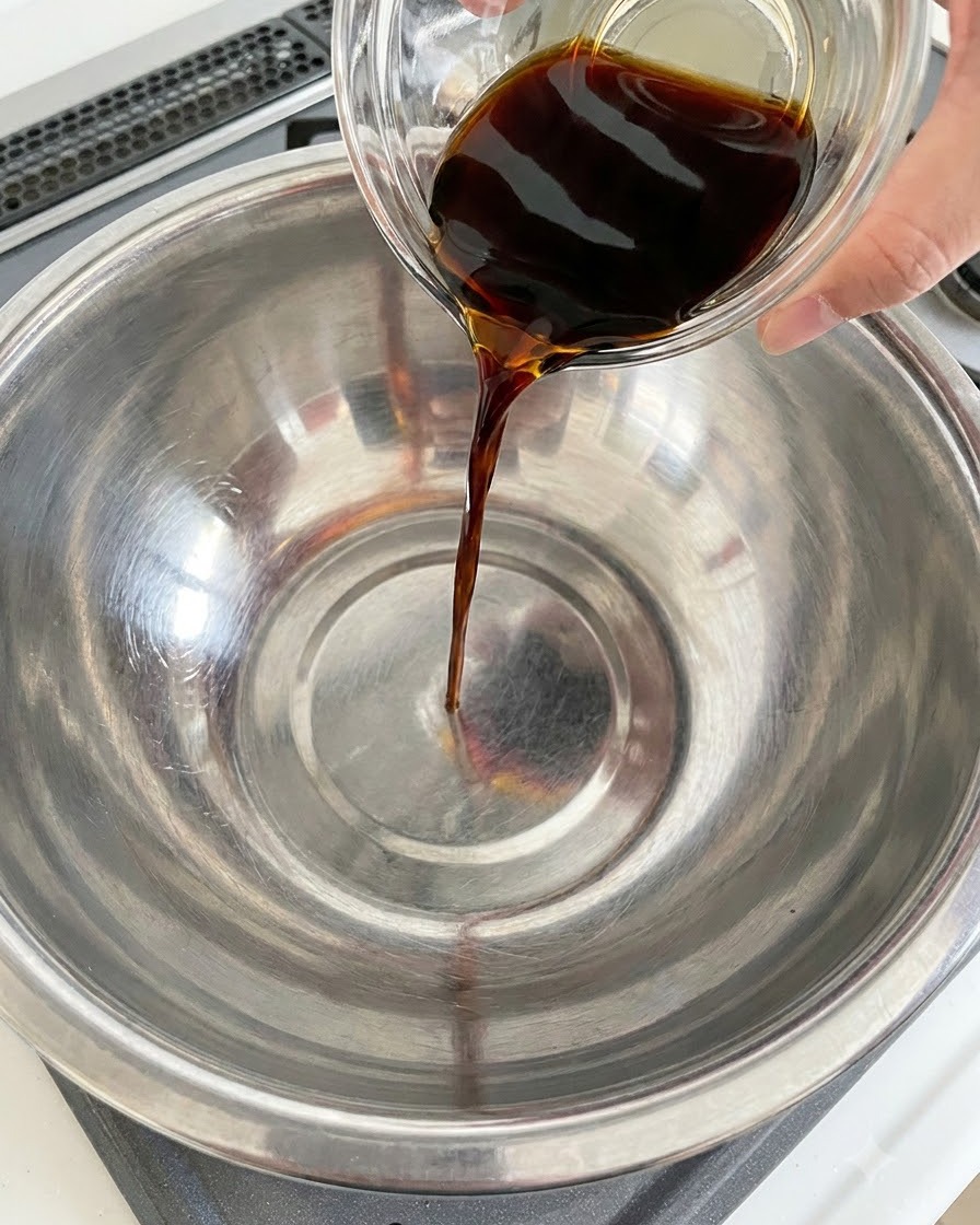 Dunkle Sojasauce wird aus einem Glasbehälter in eine Edelstahlschüssel gegossen.