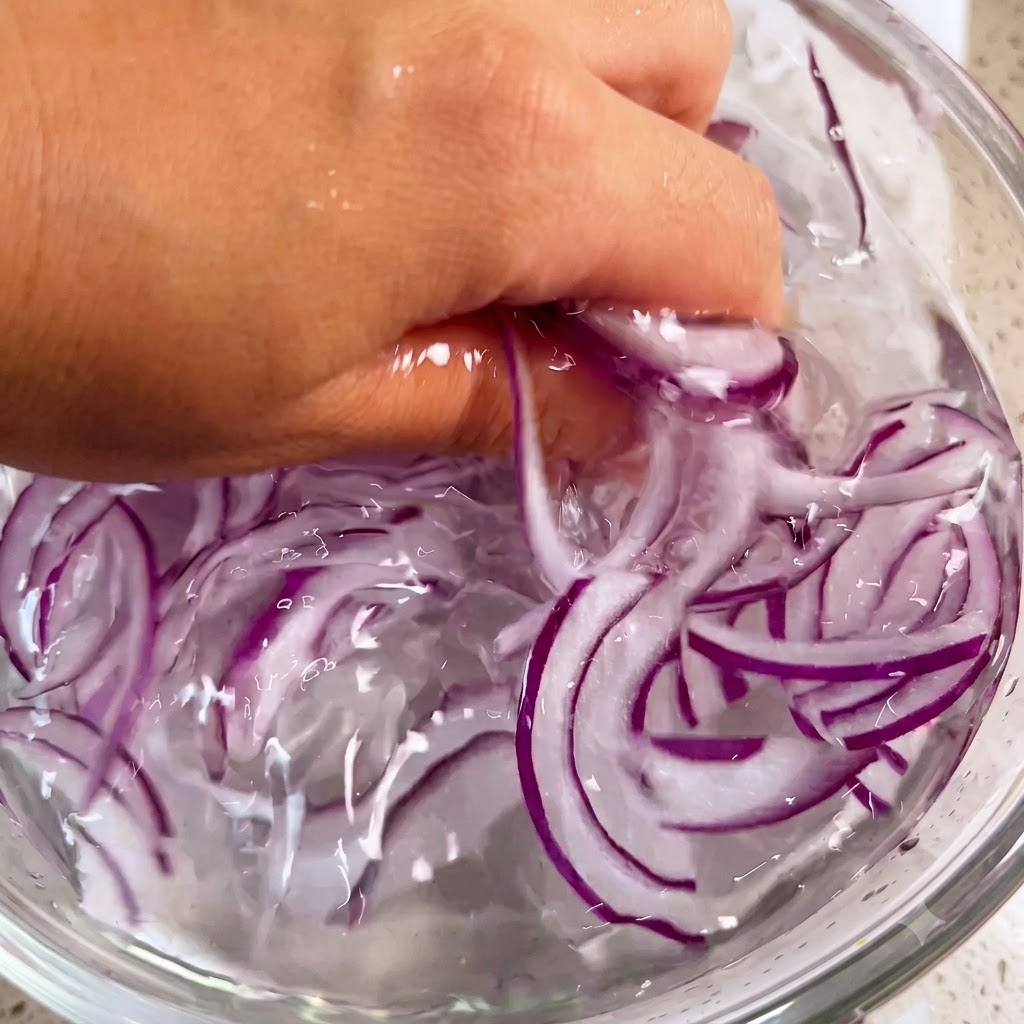 Eine Hand wäscht vorsichtig lila Zwiebelstreifen in einer Schüssel mit Eiswasser.