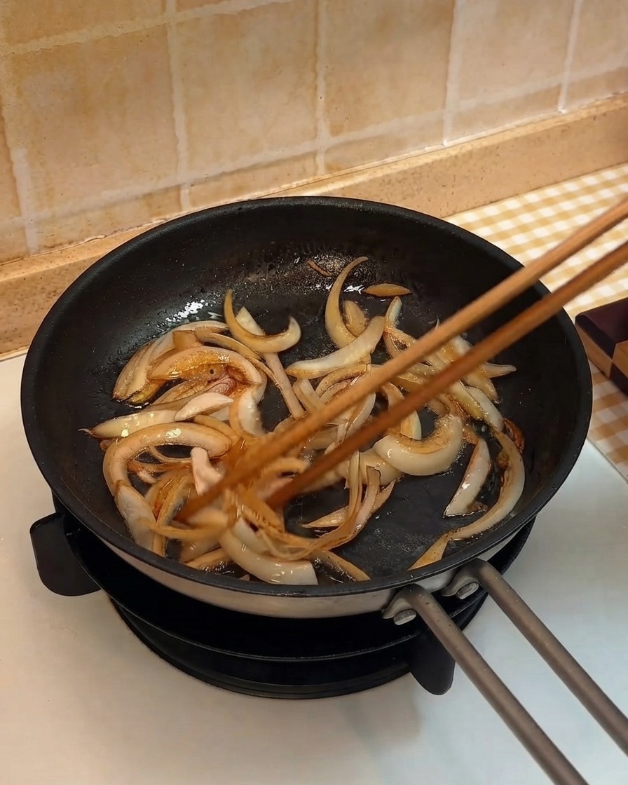 Zwiebelstreifen werden mit Holzstäbchen in einer Pfanne angebraten.