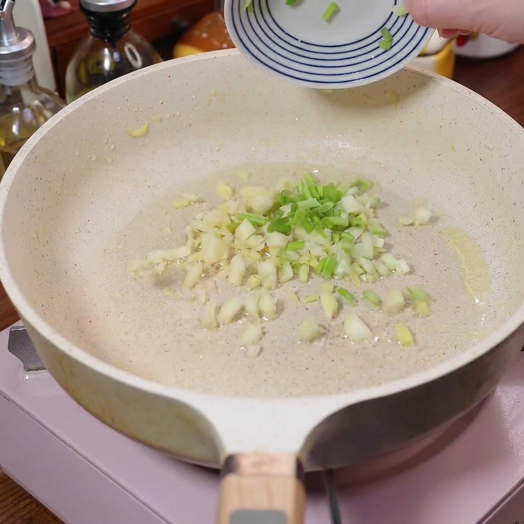 Gehackte Frühlingszwiebeln, Knoblauch und Ingwer brutzeln in einer weißen Pfanne.