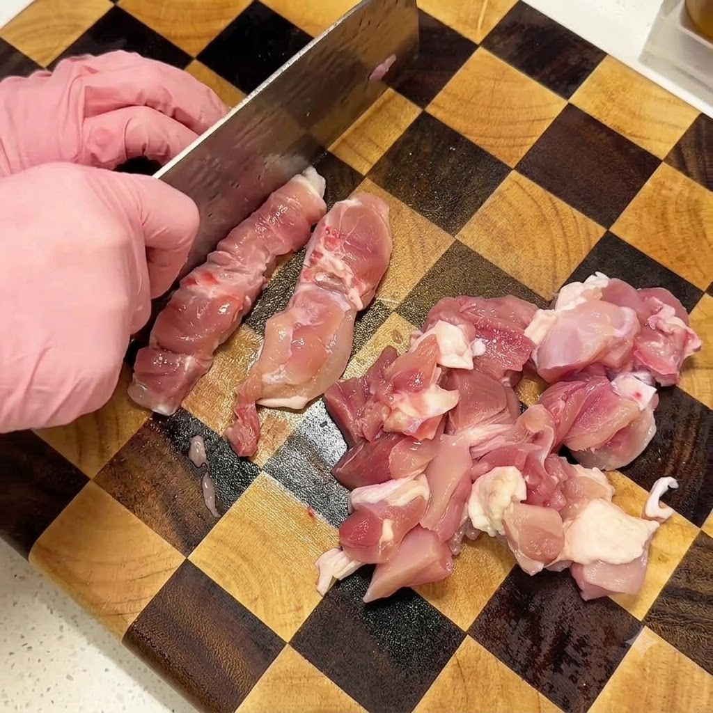Hände in rosa Handschuhen schneiden rohe Hähnchenschenkel mit einem Hackbeil auf einem karierten Holzschneidebrett in Würfel.