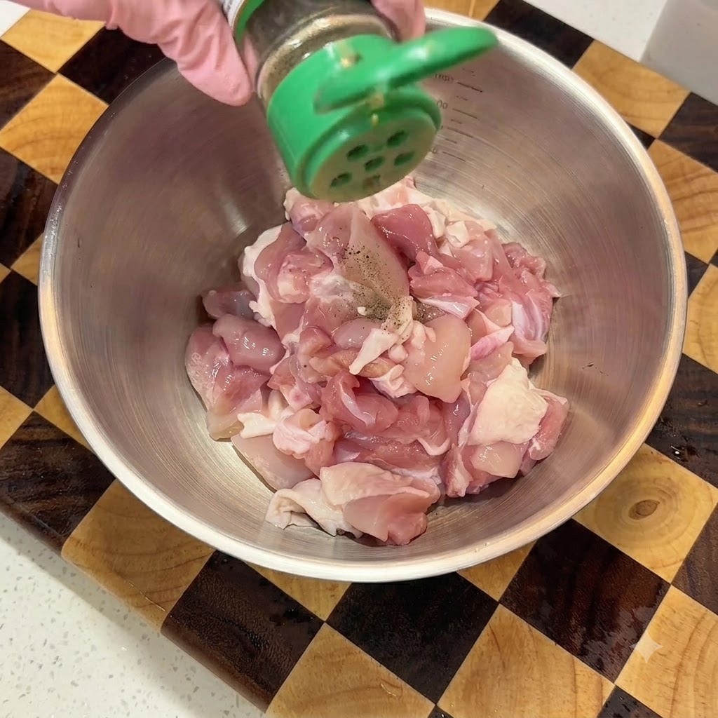 Schwarzes Pfefferpulver wird aus einem grünen Streuer auf gewürfeltes rohes Hähnchenfleisch in einer Metallschüssel gestreut.