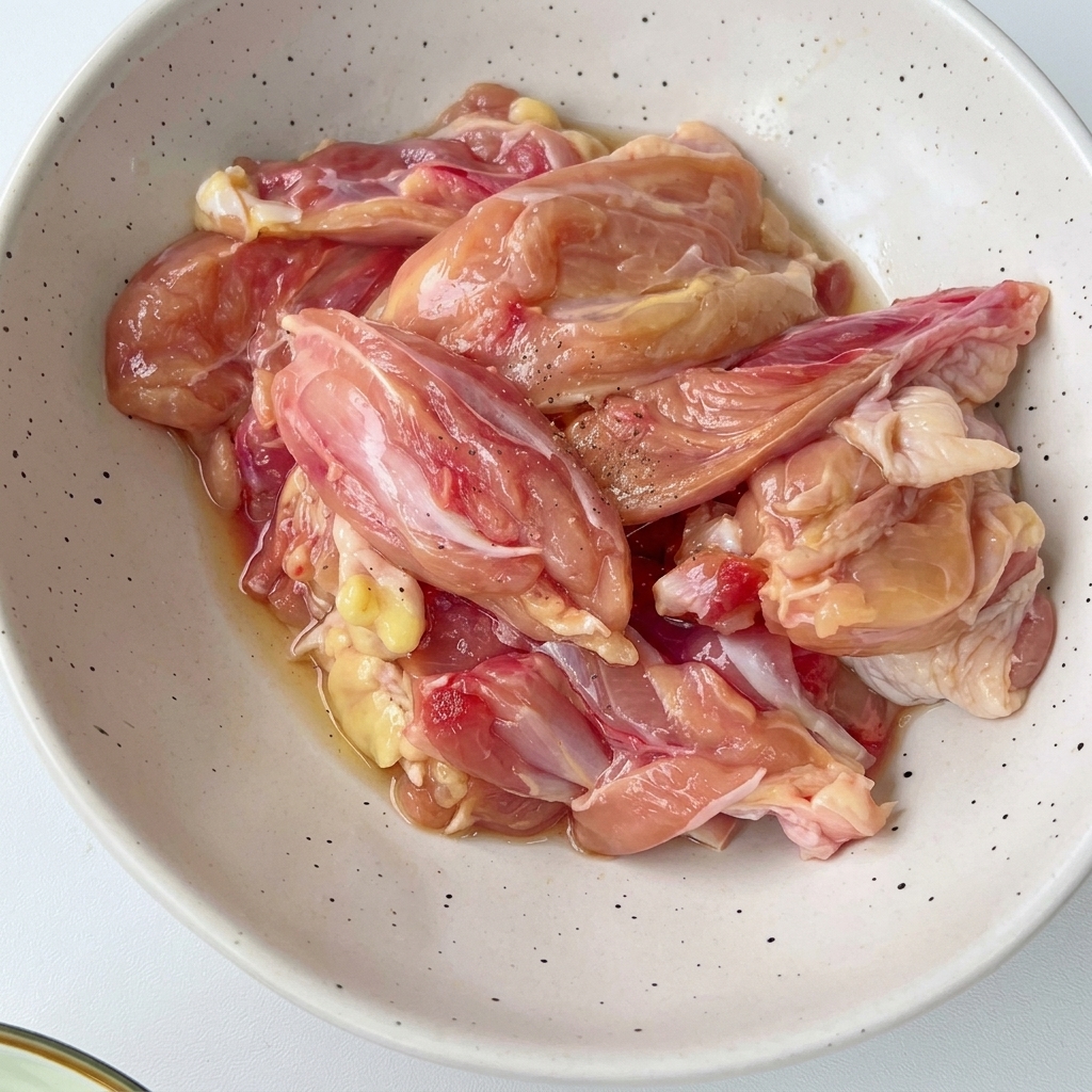 Geschnittene Hähnchenstücke in einer weiß gesprenkelten Schüssel, vermischt mit Gewürzen und Aromen.