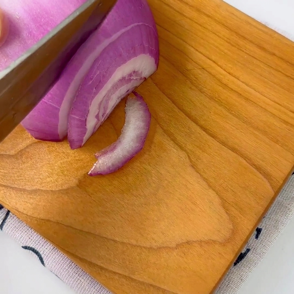 Eine rote Zwiebel wird mit einem Messer auf einem Holzschneidebrett in dünne Streifen geschnitten.