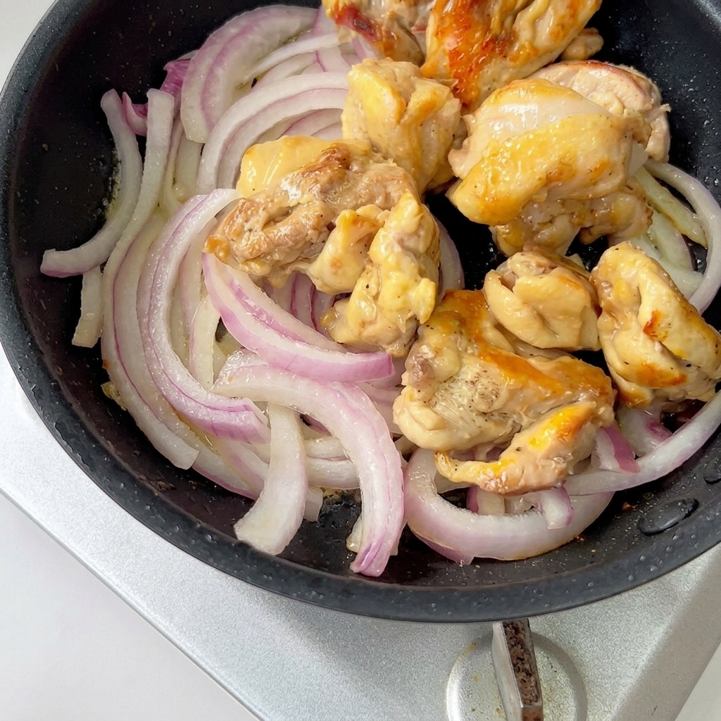 Gekochte Hähnchenstücke und geschnittene rote Zwiebeln köcheln in einer Pfanne mit Sauce.