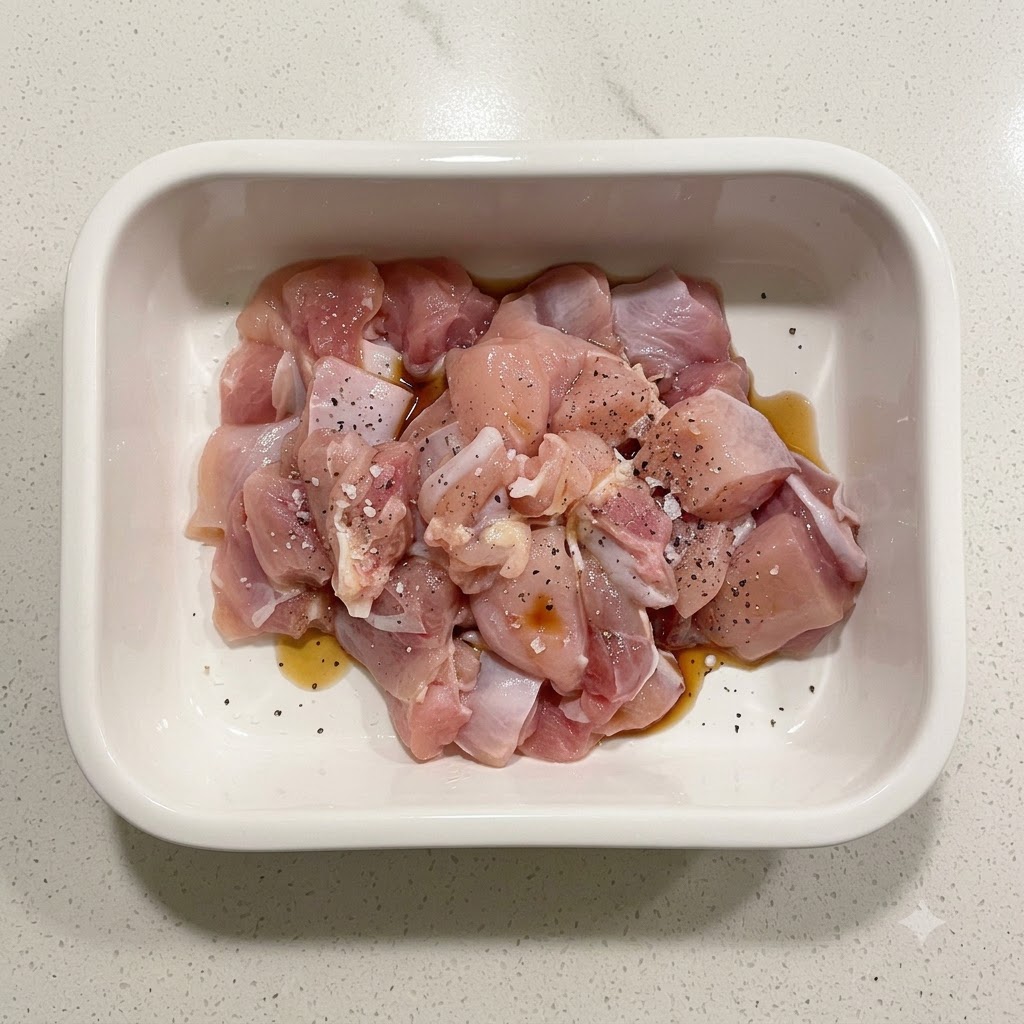 Rohe Hähnchenstücke, gewürzt mit Stärke, Sojasoße und schwarzem Pfeffer in einem weißen Keramikbehälter.